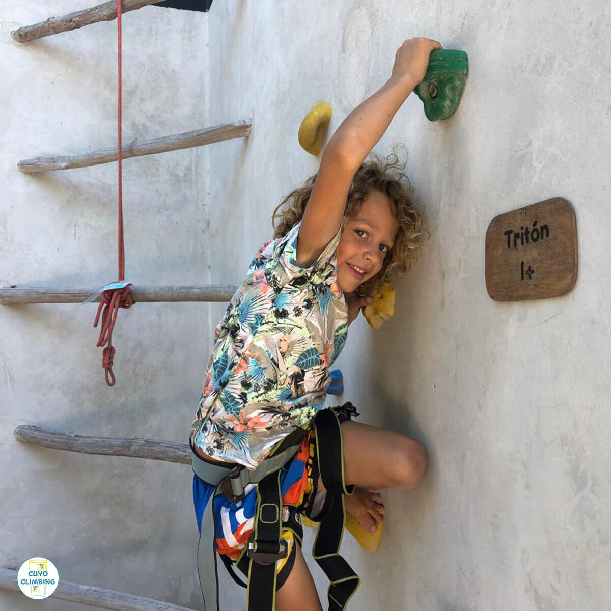 Escalada para niños en Cuyo Climbing Un a niño con arnés alcanza un hold verde mientras escala el muro.