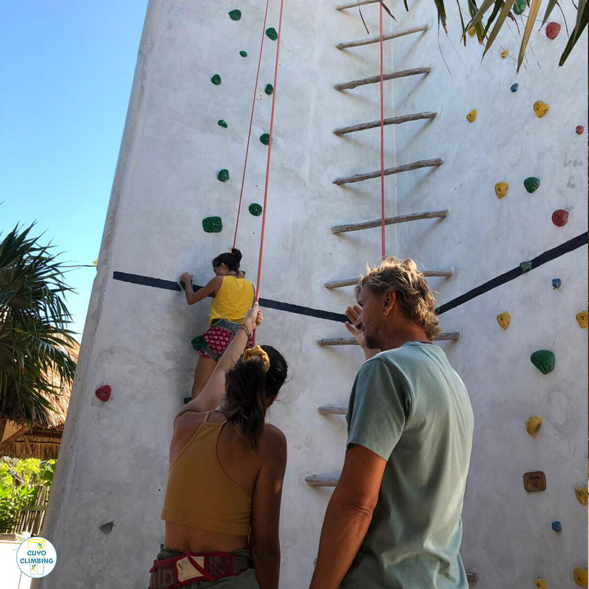 Un instructor guía a una participante durante el curso de iniciación, mientras otra persona escala el muro al aire libre.