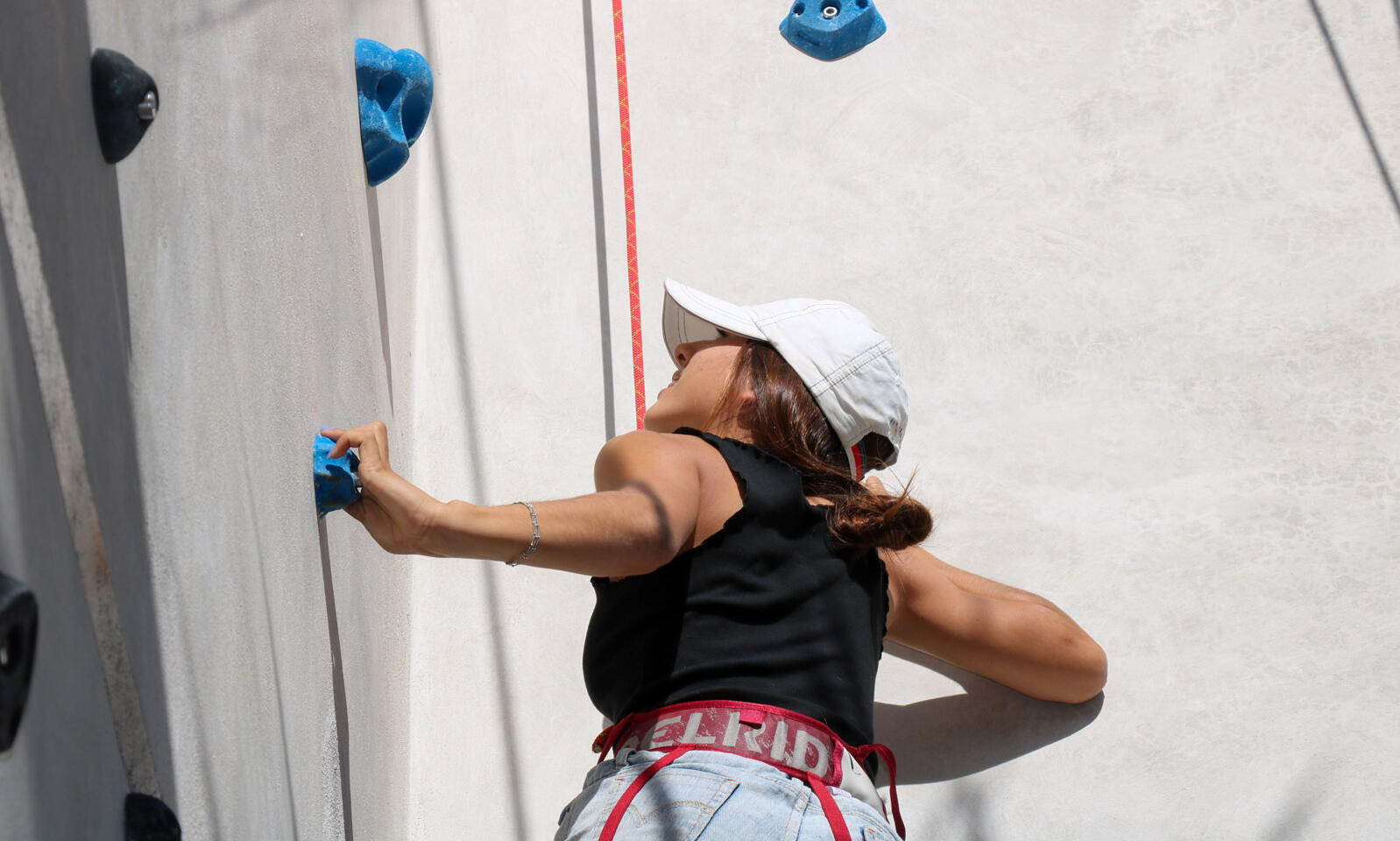 Una escaladora con gorra blanca y arnés alcanza una presa azul en el muro de Cuyo Climbing.